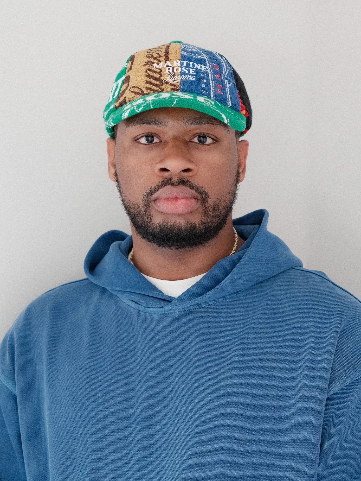 A young man wearing a colorful patchwork baseball cap and a blue hoodie, standing against a plain light gray background.