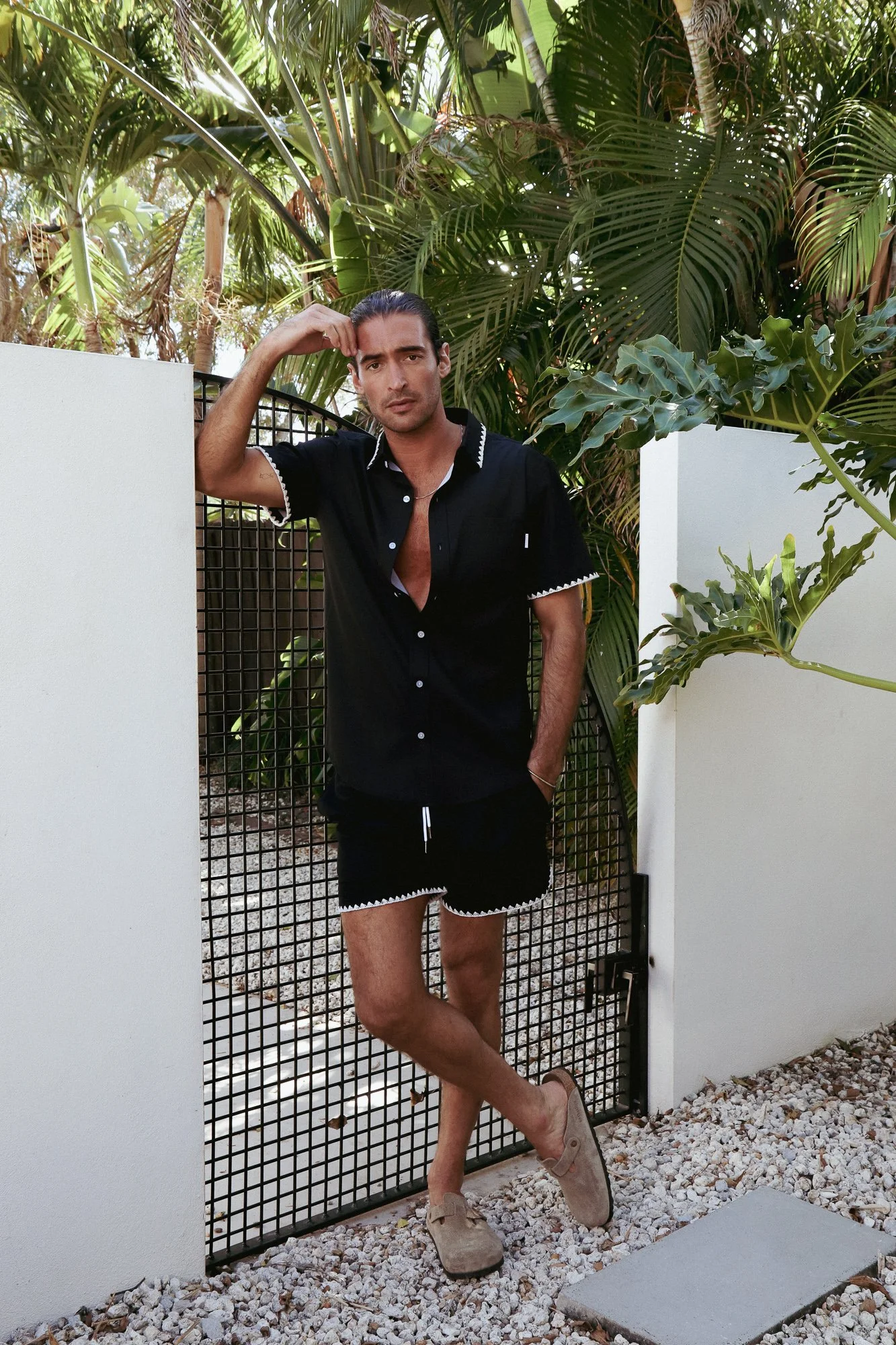 A man standing outdoors with lush tropical greenery behind him, wearing a black short-sleeve shirt, black shorts, tan boots, and sunglasses, with one hand on his head.
