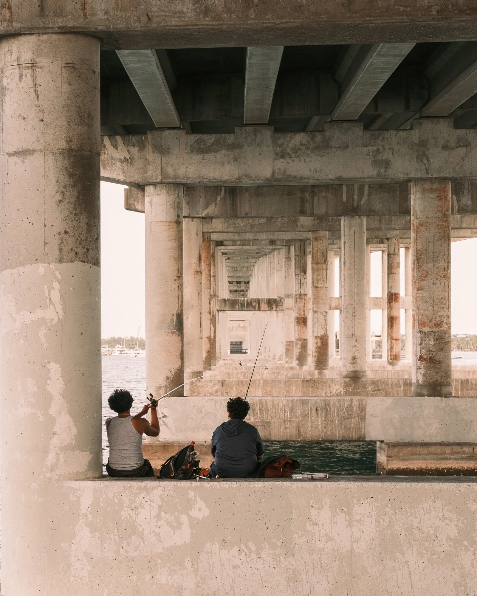 Two men fishing under a bridge over water, with a scenic view of boats and the horizon in the background.