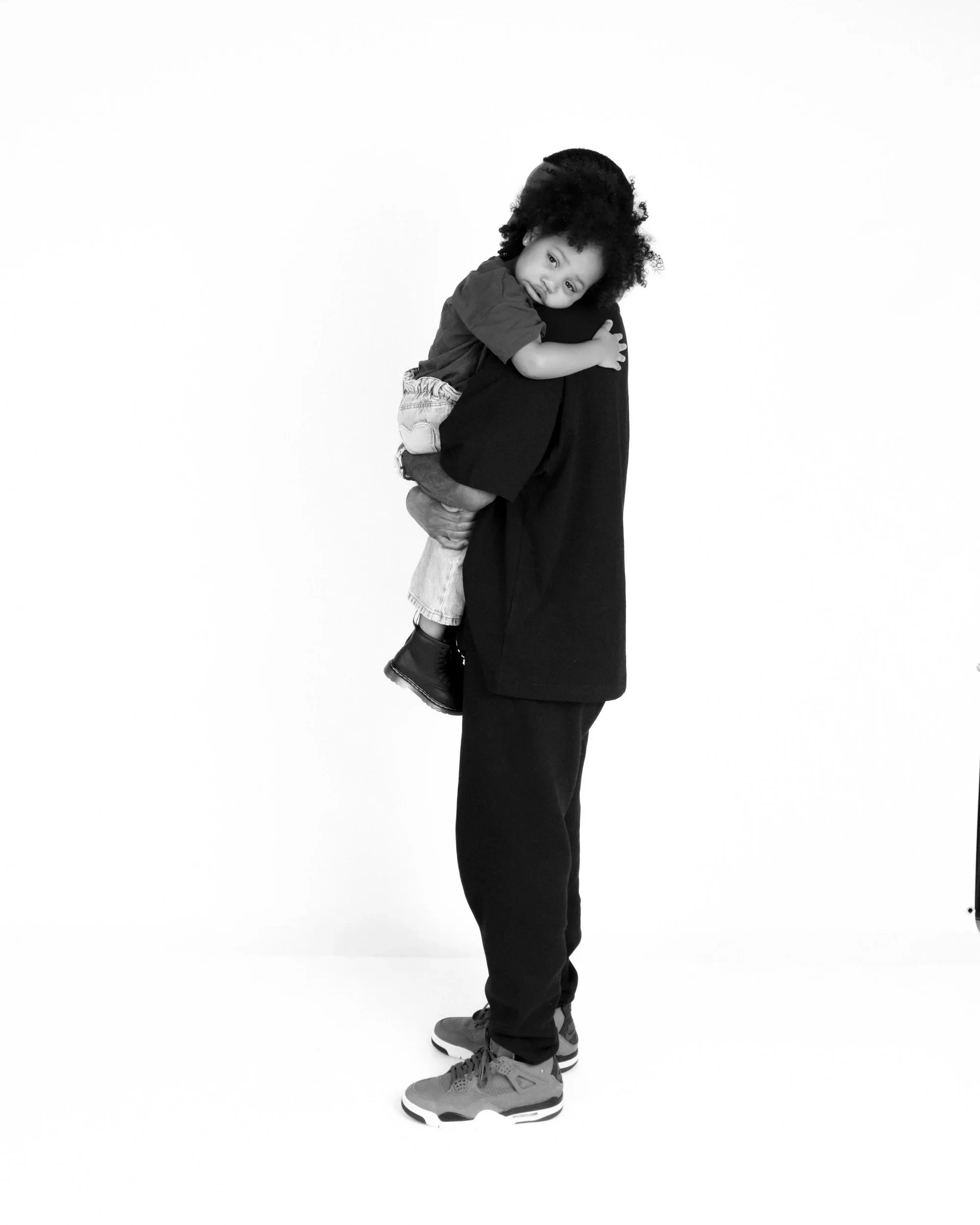 A person wearing black clothing and Nike sneakers holding a young child with curly hair. The child is resting their head on the person's shoulder and looking at the camera with a gentle expression. The background is plain white.
