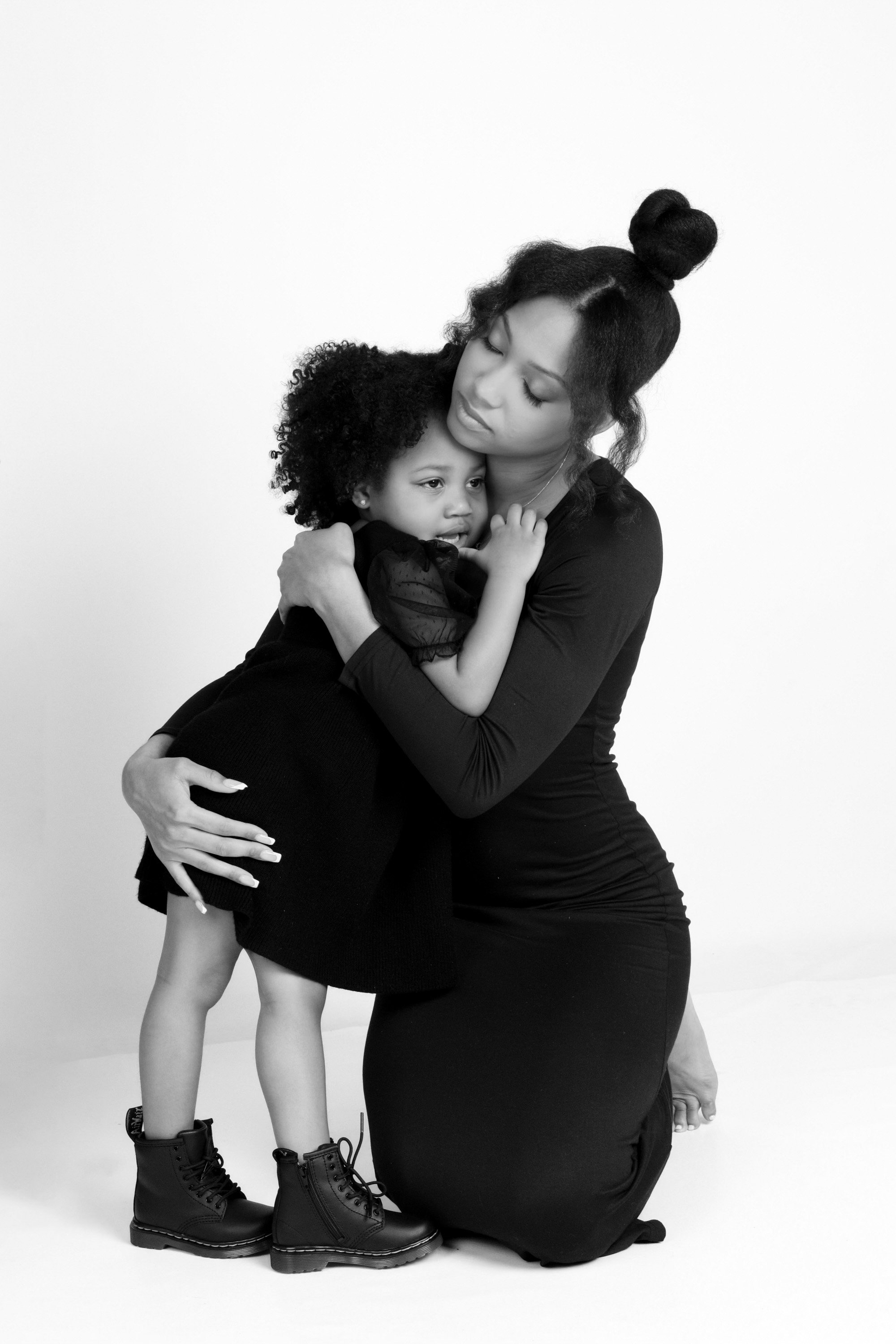 A woman kneeling and hugging a young girl, both with dark curly hair, against a plain white background. The girl is wearing black boots and a black dress, and the woman is dressed in a long black dress.
