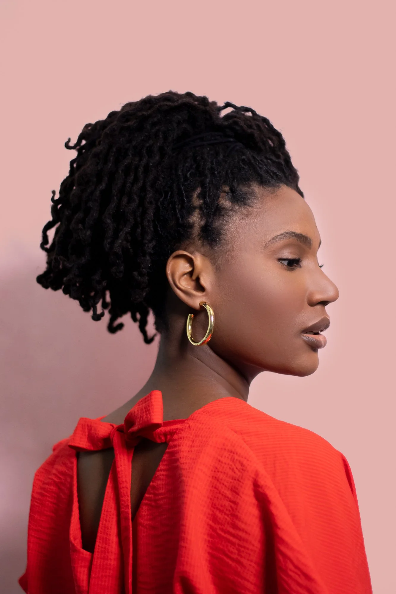 A profile view of a woman with dark skin and dreadlocks tied up in an elegant style, wearing a red dress with a back tie, large gold hoop earrings, and a confident expression against a pale pink background.