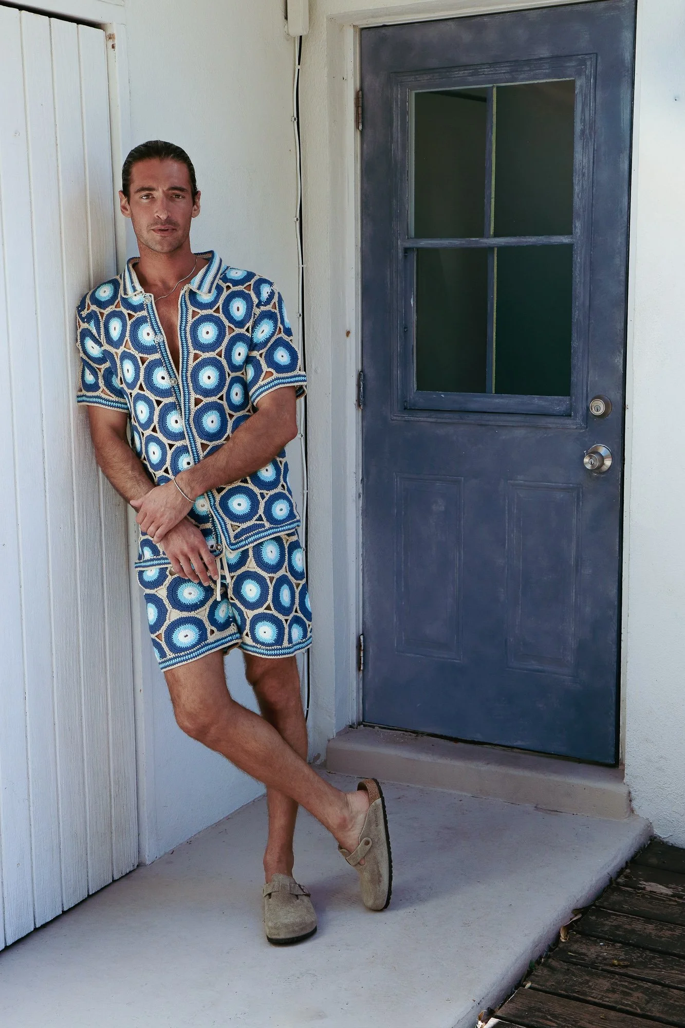 A man with dark hair and a light complexion is standing outside a house next to a blue front door. He is wearing a matching blue, white, and brown patterned shirt and shorts, tan slip-on shoes, and is holding a glass of drink. He is leaning with his 