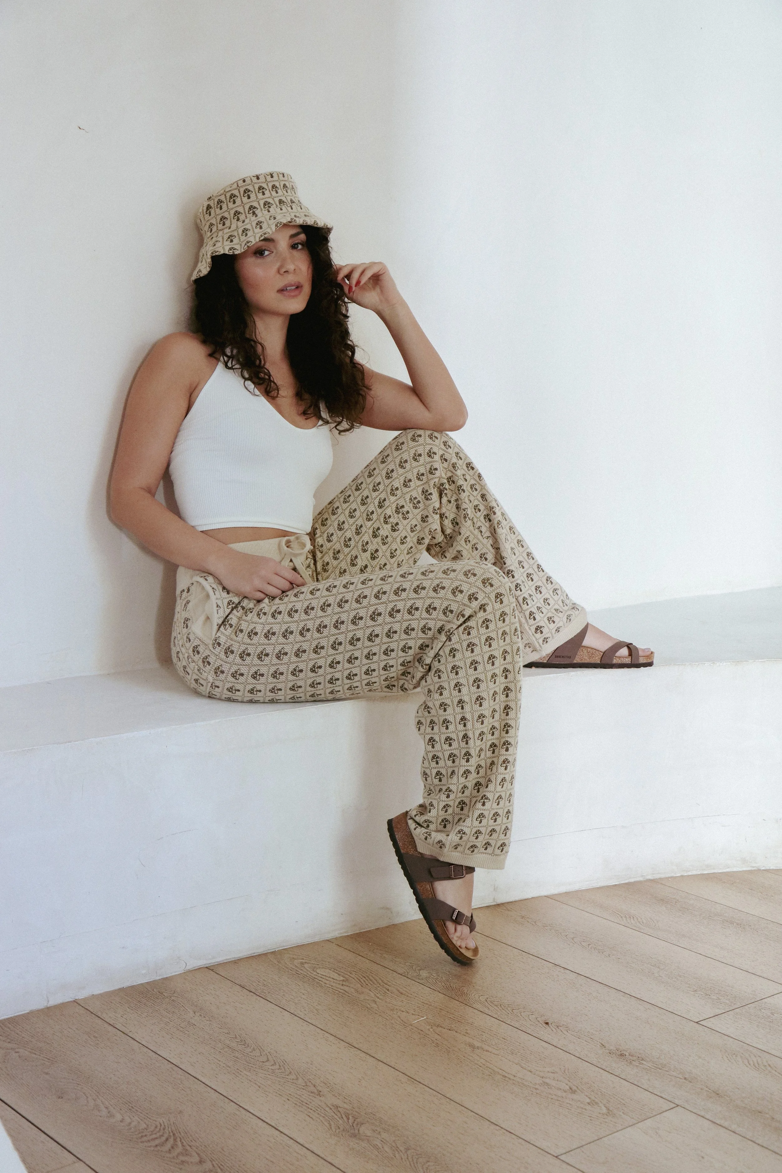A woman sits on a white ledge against a plain white wall, wearing a beige patterned bucket hat, a white tank top, beige patterned wide-leg pants, and brown sandals. She has dark curly hair and is looking at the camera. The floor is light wood.