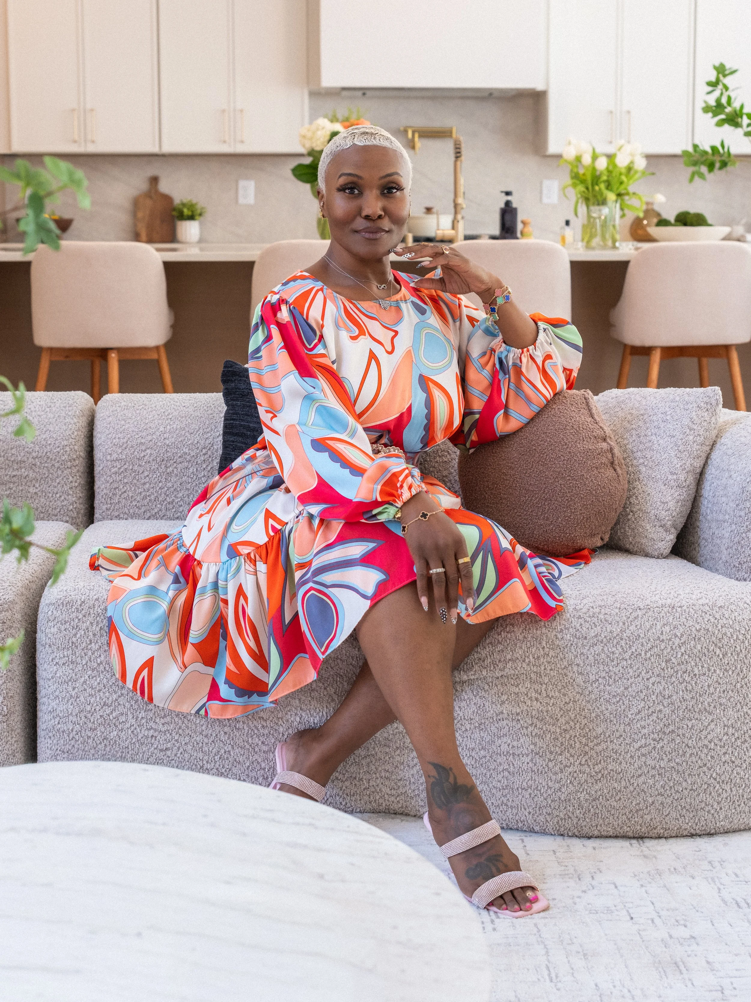 A woman with short platinum blonde hair sitting on a beige couch in a modern kitchen, wearing a colorful patterned dress, smiling softly, with flowers and greenery in the background.