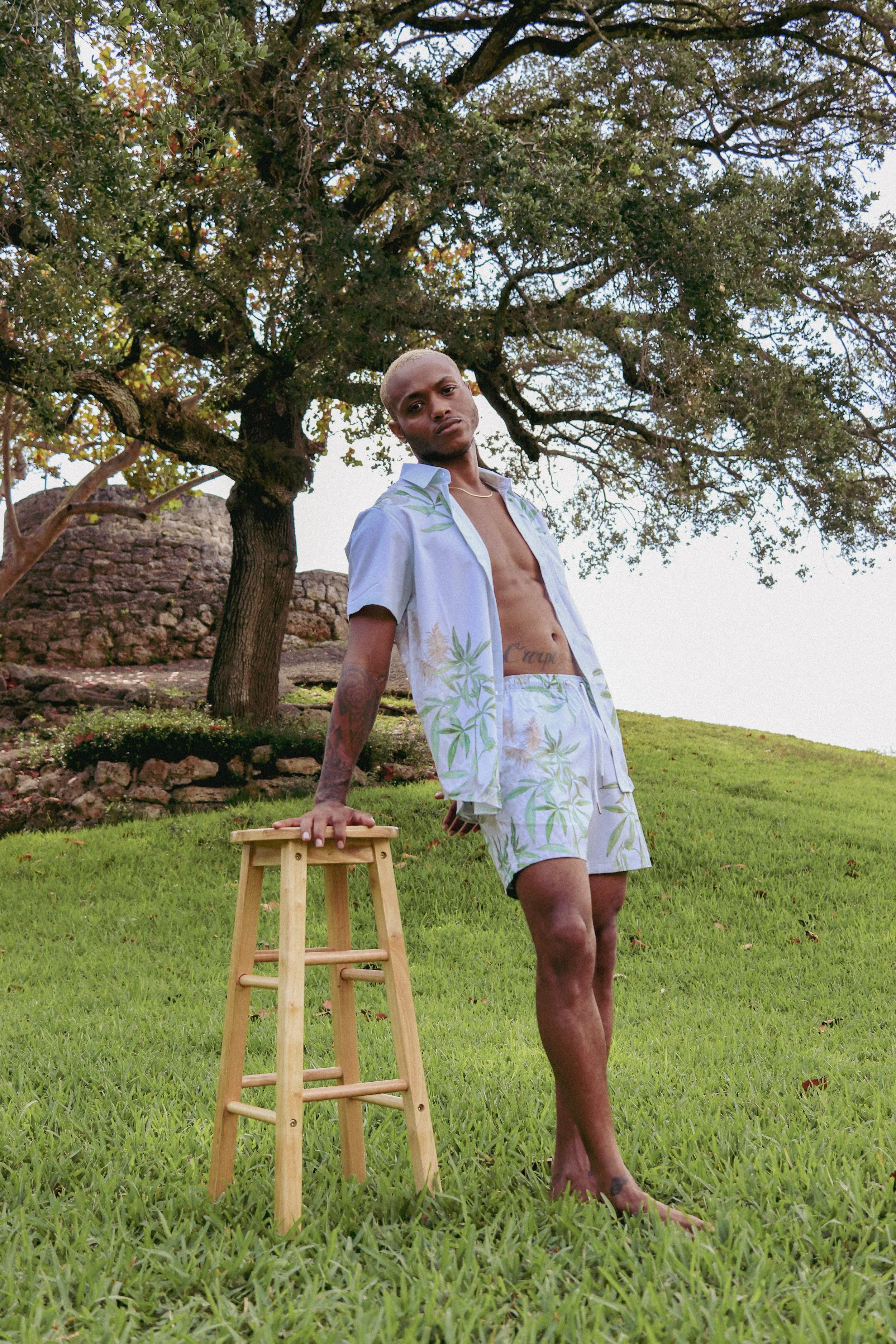A young man with blond hair and tattoos stands outdoors on a grassy lawn, leaning on a small wooden stool with one hand. He wears a light blue shirt with green leaf patterns, unbuttoned to reveal his chest, and matching shorts. Behind him, there is a