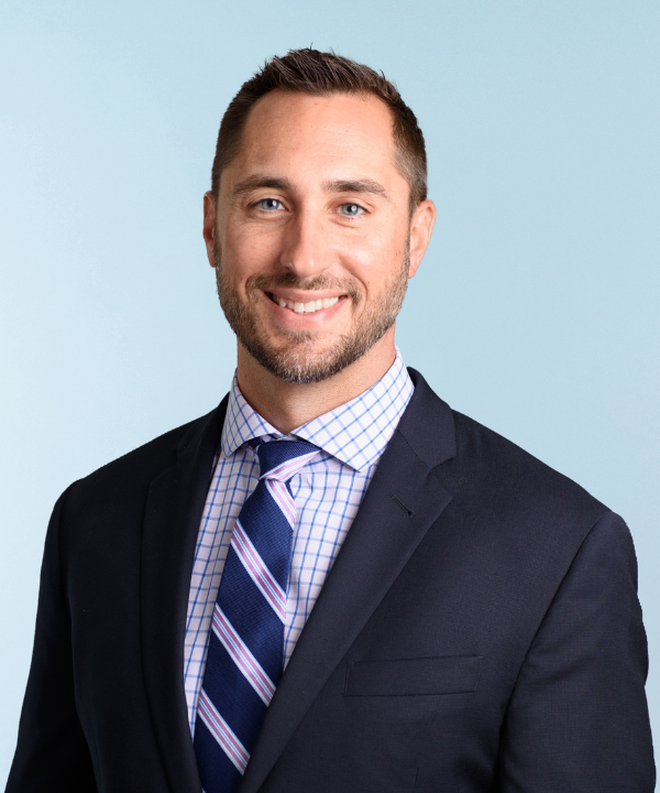 A smiling man in a dark suit, checkered shirt, and striped tie posing against a light blue background.