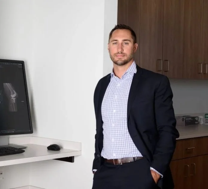 A man in business attire standing in an office or medical setting, with medical imaging on a computer monitor behind him.