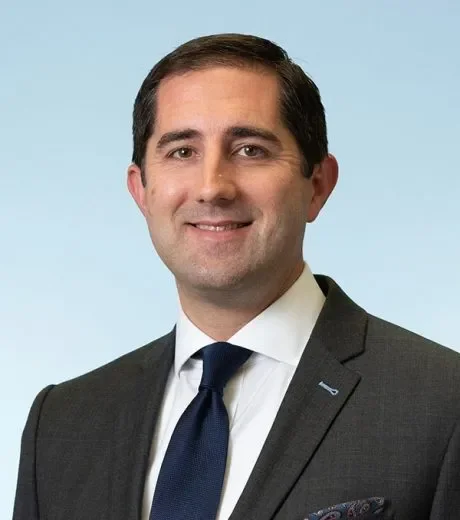 A man with dark brown hair, wearing a dark suit, white shirt, and navy blue tie, smiling against a light blue background.