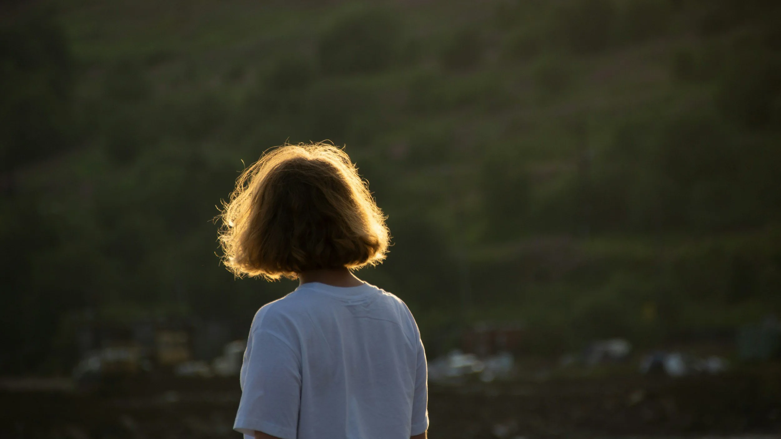 Back view of a person with shoulder-length blonde hair wearing a white shirt, standing outdoors during sunset or sunrise.