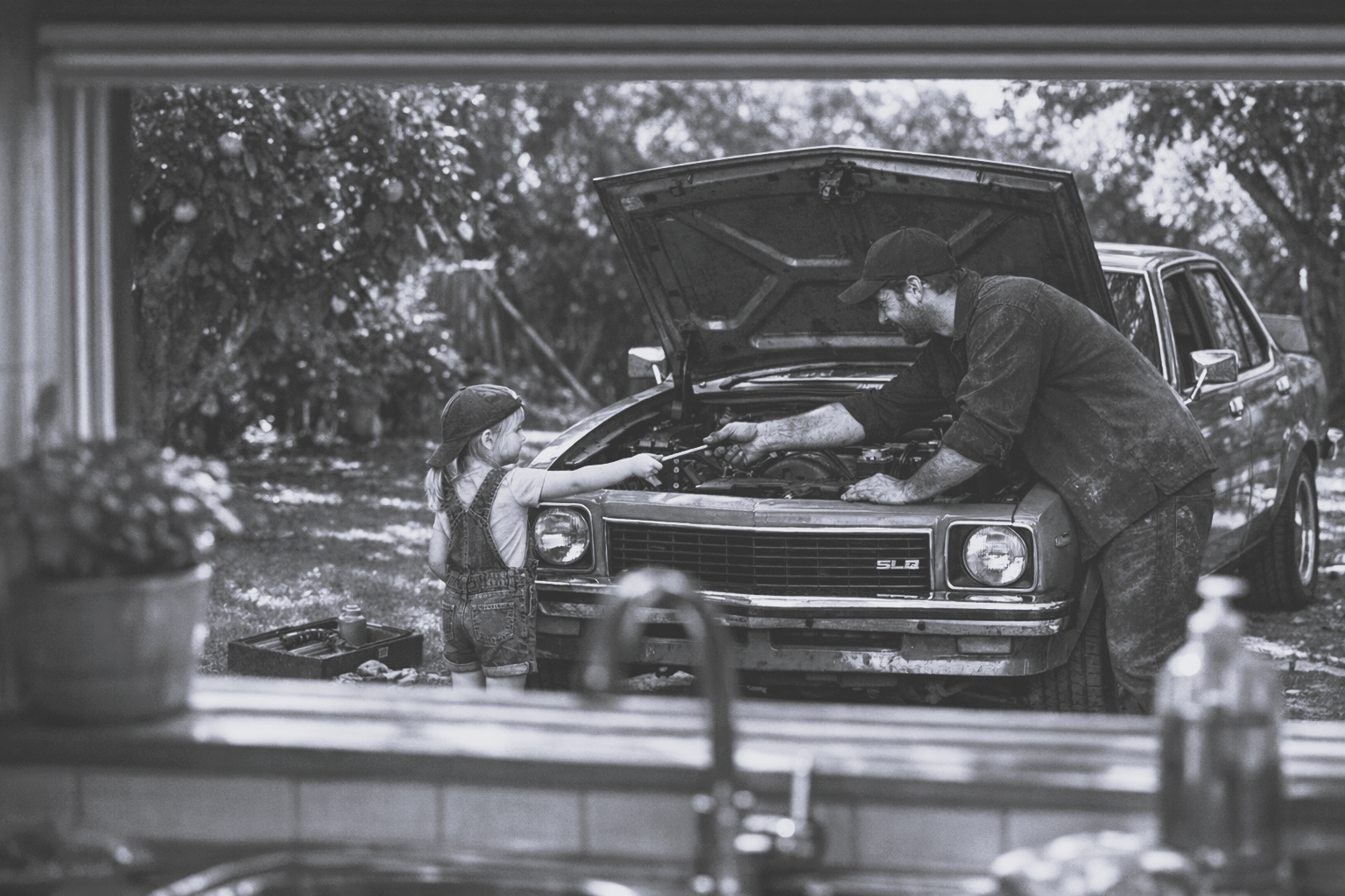 A black and white image of a daughter helping fix a car engine, handing a tool to her father.
