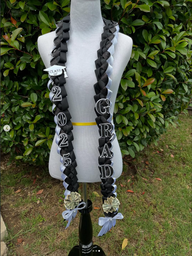 Graduation leis with black and white ribbons, foam dollar bills, and decorative letters spelling 'GRADED 2025' on a mannequin outside among green foliage.