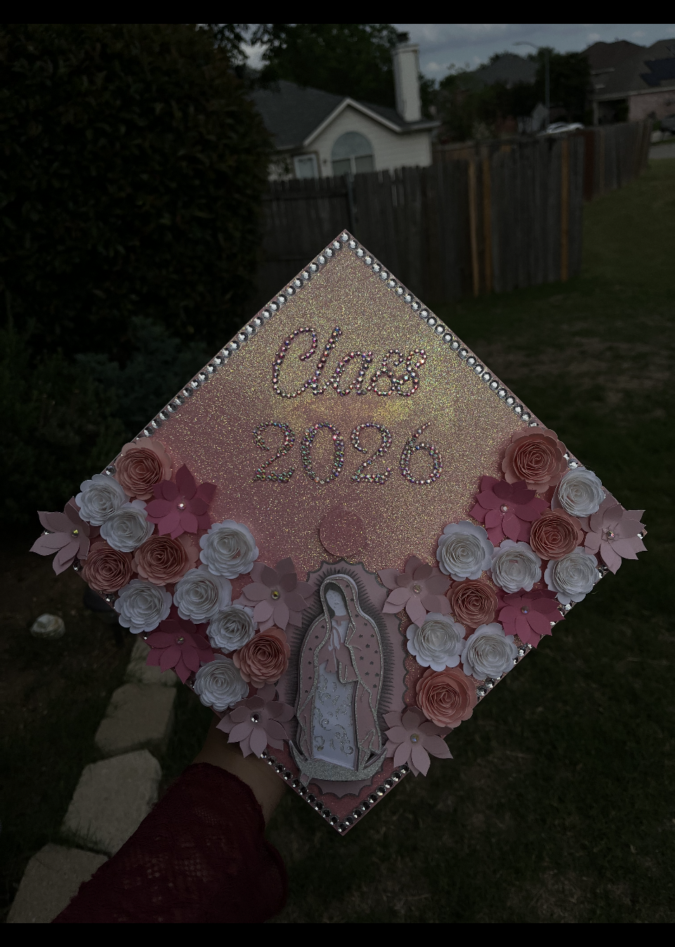 Decorated graduation cap with pink, white, and peach paper flowers, the phrase "Claus 2020" in rhinestones, and an image of Jesus Christ, held outside in a backyard.