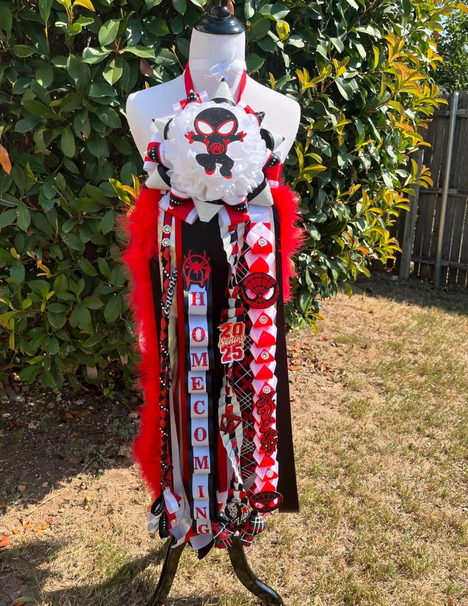 A decorated mannequin outdoors with a hedge and fence in the background, adorned with various ribbons, ornaments, and decorations celebrating a child's first grade and birthday, featuring a festive color scheme of red, black, and white.