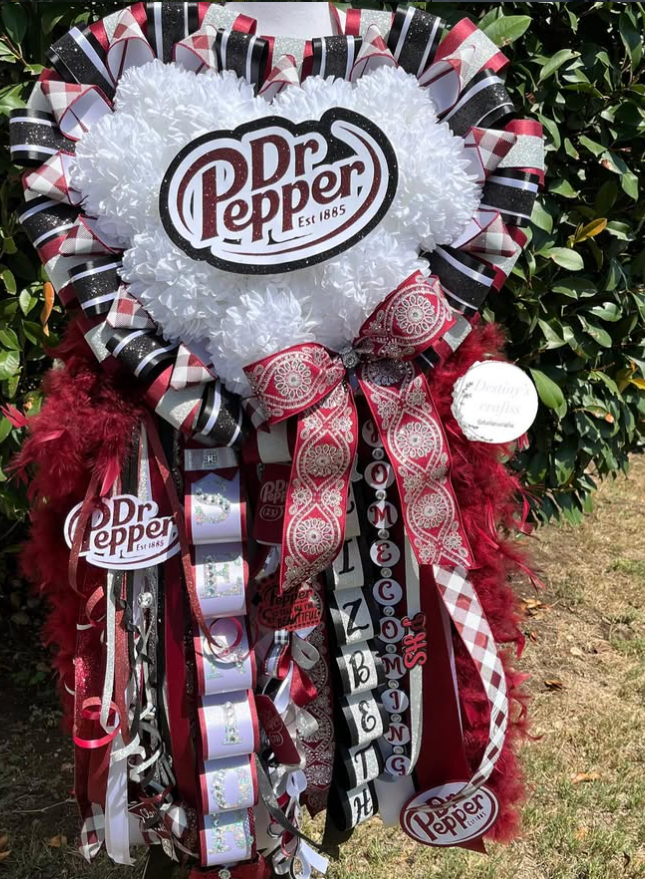 A decorative wreath with Dr. Pepper logos, ribbons, and bows, placed outdoors among green bushes.
