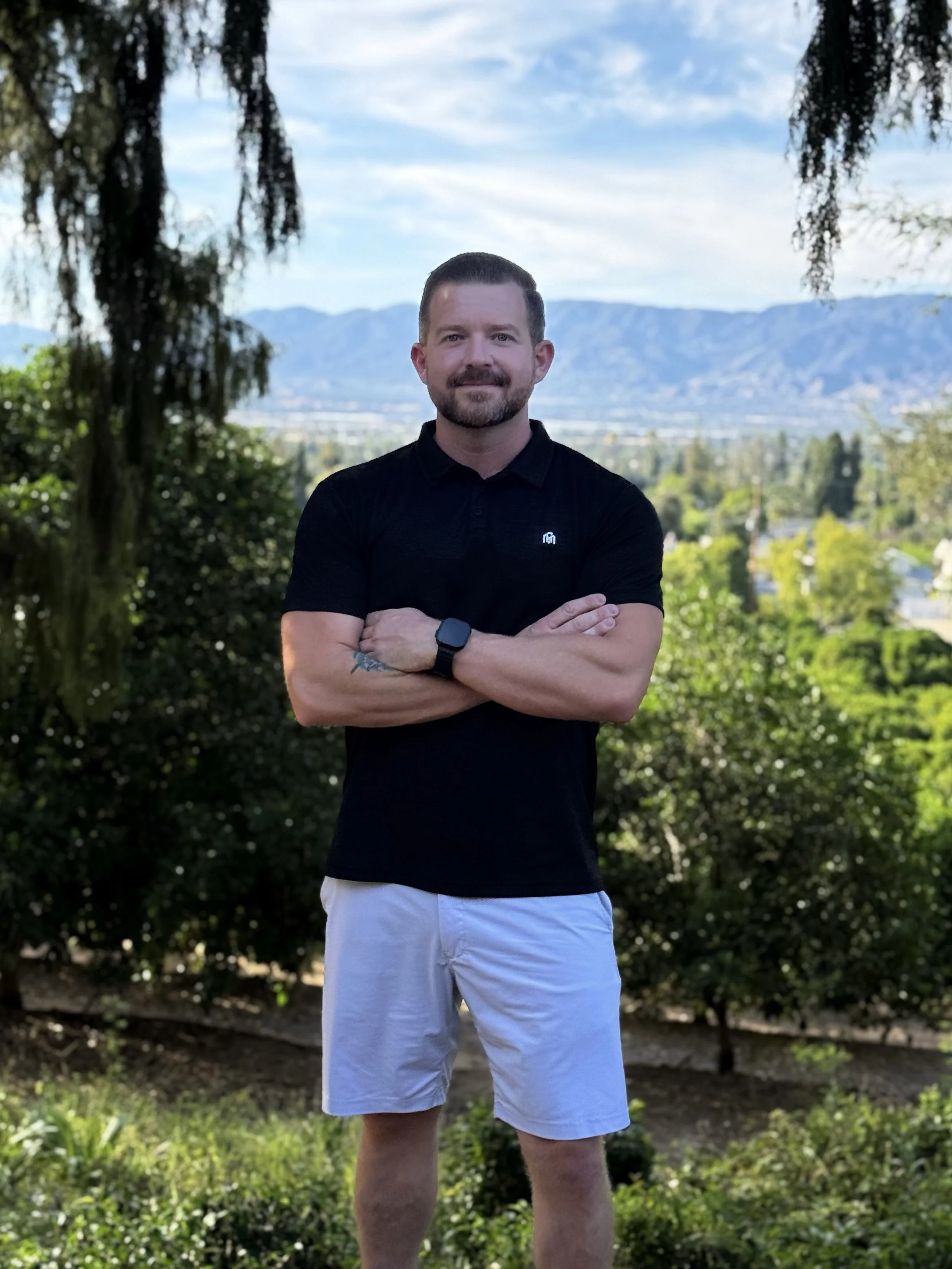 A man standing outdoors in a park with trees and mountains in the background. He has a beard, short hair, and is wearing a black polo shirt, white shorts, and a smartwatch. He is crossing his arms and smiling at the camera.
