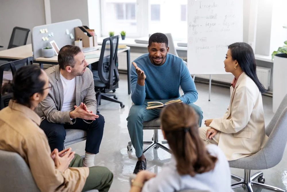 Group of diverse coworkers having a discussion in a modern office conference room, with a man in blue speaking and others listening.