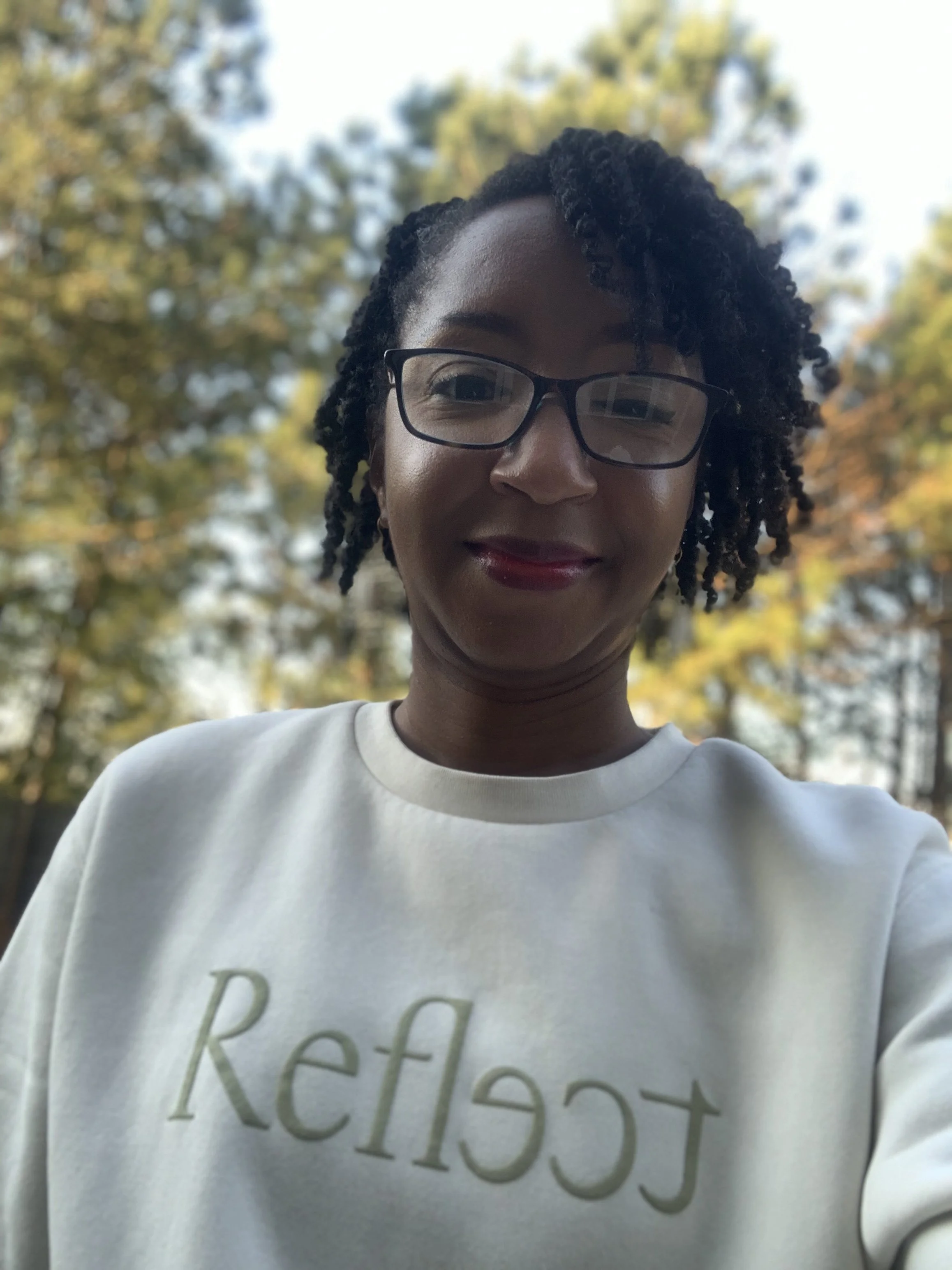 A woman with glasses and short curly hair taking a selfie outdoors with trees and blue sky in the background.