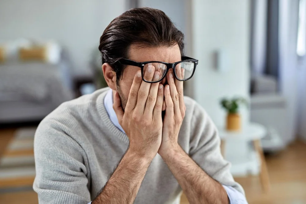 Man with glasses covering his face with both hands in a living room