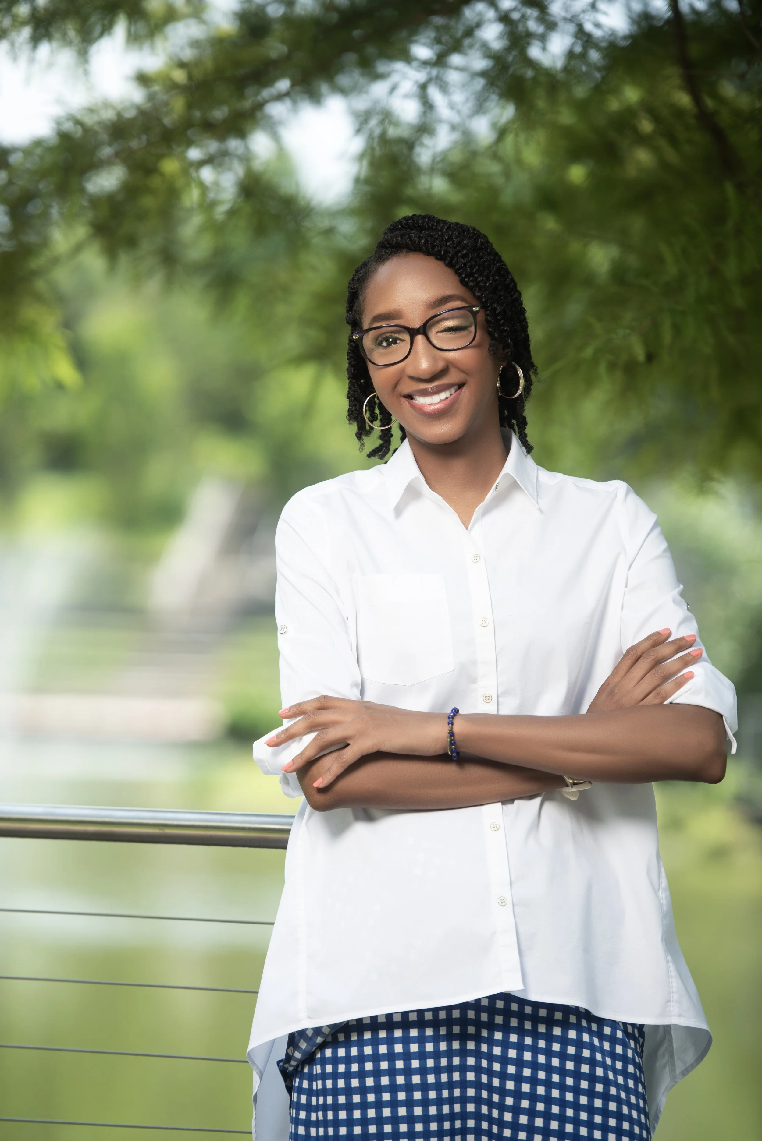 Smiling woman with glasses and earrings outdoors, wearing a white blouse and patterned skirt, arms crossed, with green trees in the background.
