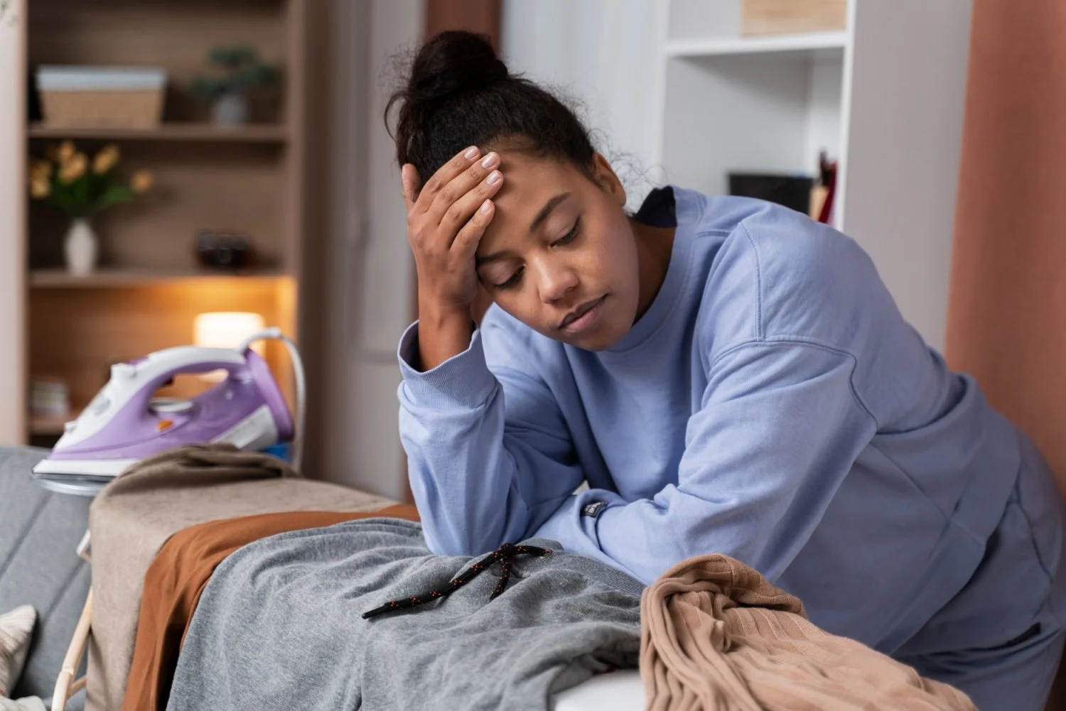 Woman with a worried expression, holding her forehead, sitting next to an ironing board and clothes.