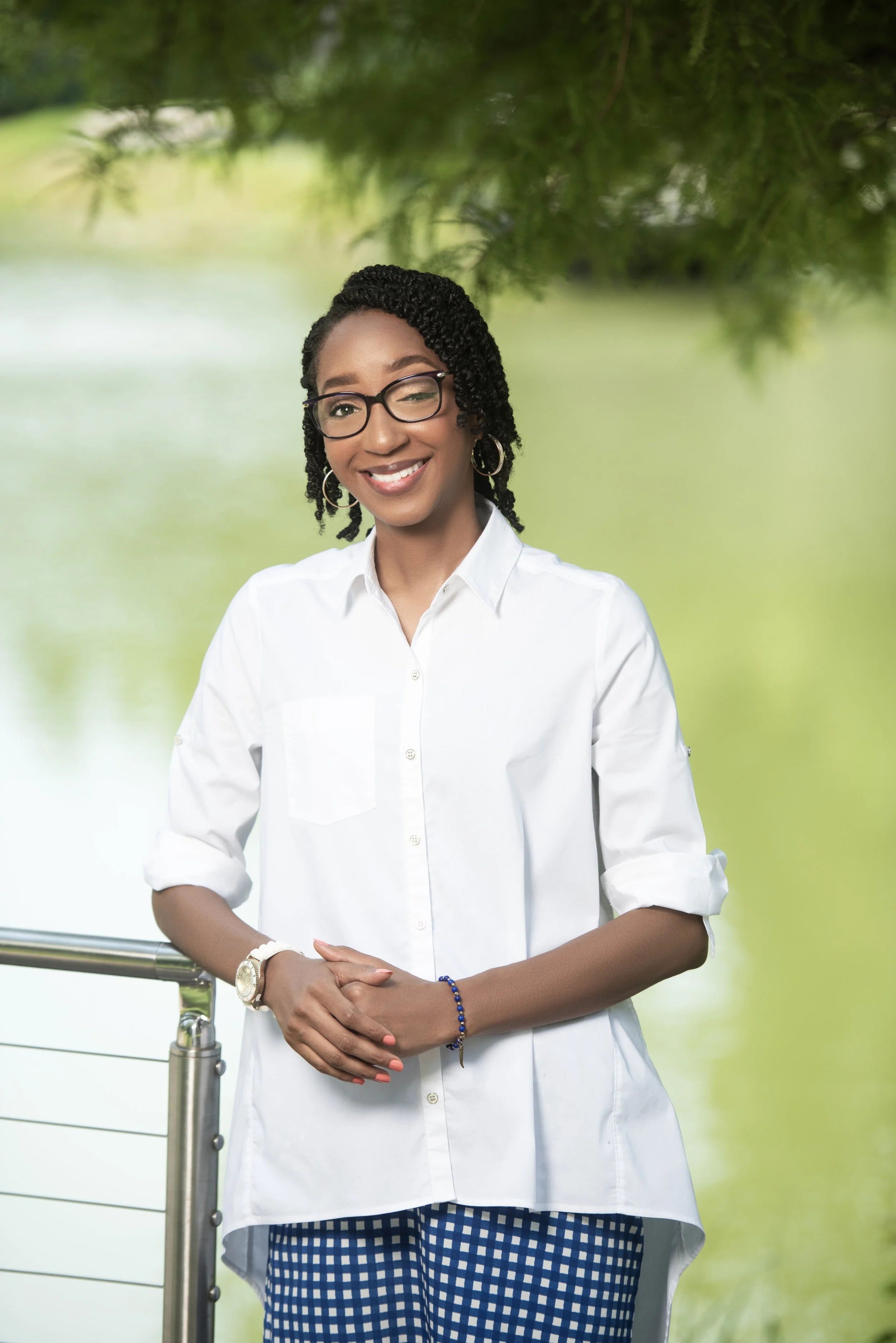 A smiling woman with glasses and braided hair, wearing a white shirt and blue checkered pants, stands outdoors near a railing with greenery in the background.