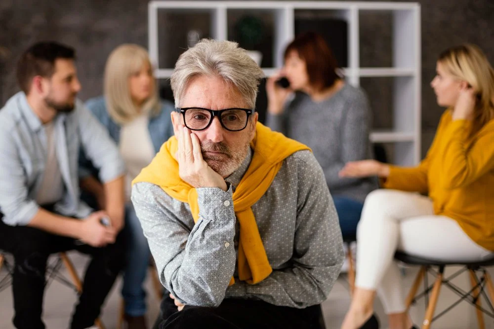 Man with gray hair and glasses looking bored or frustrated at a group therapy session.
