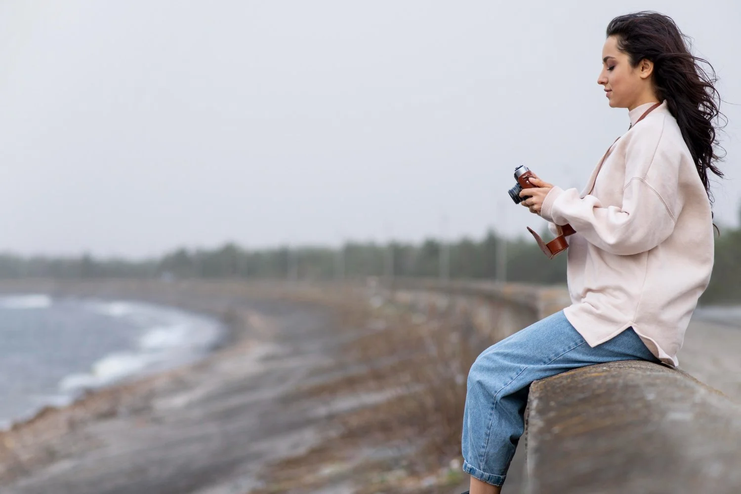 Young woman sitting on a concrete barrier by the ocean, holding a vintage camera and looking down.
