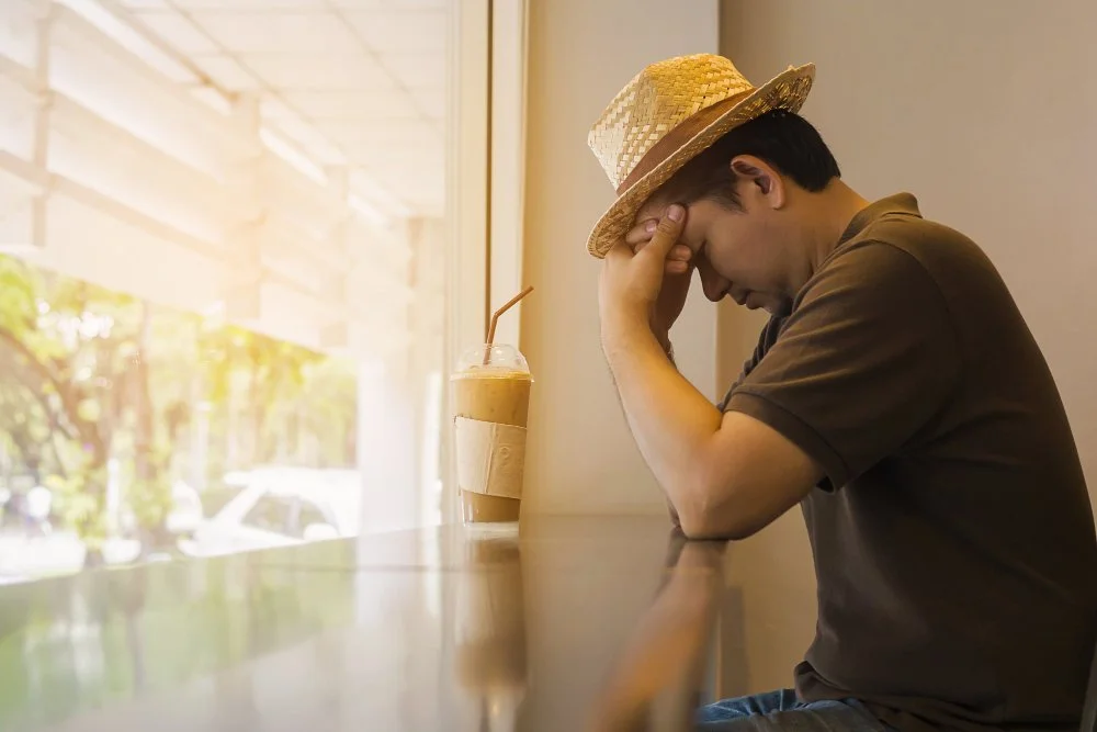 A man wearing a straw hat sitting at a cafe table with his head resting on his hand, looking down and appearing upset. There is a beverage with a straw on the table beside him.