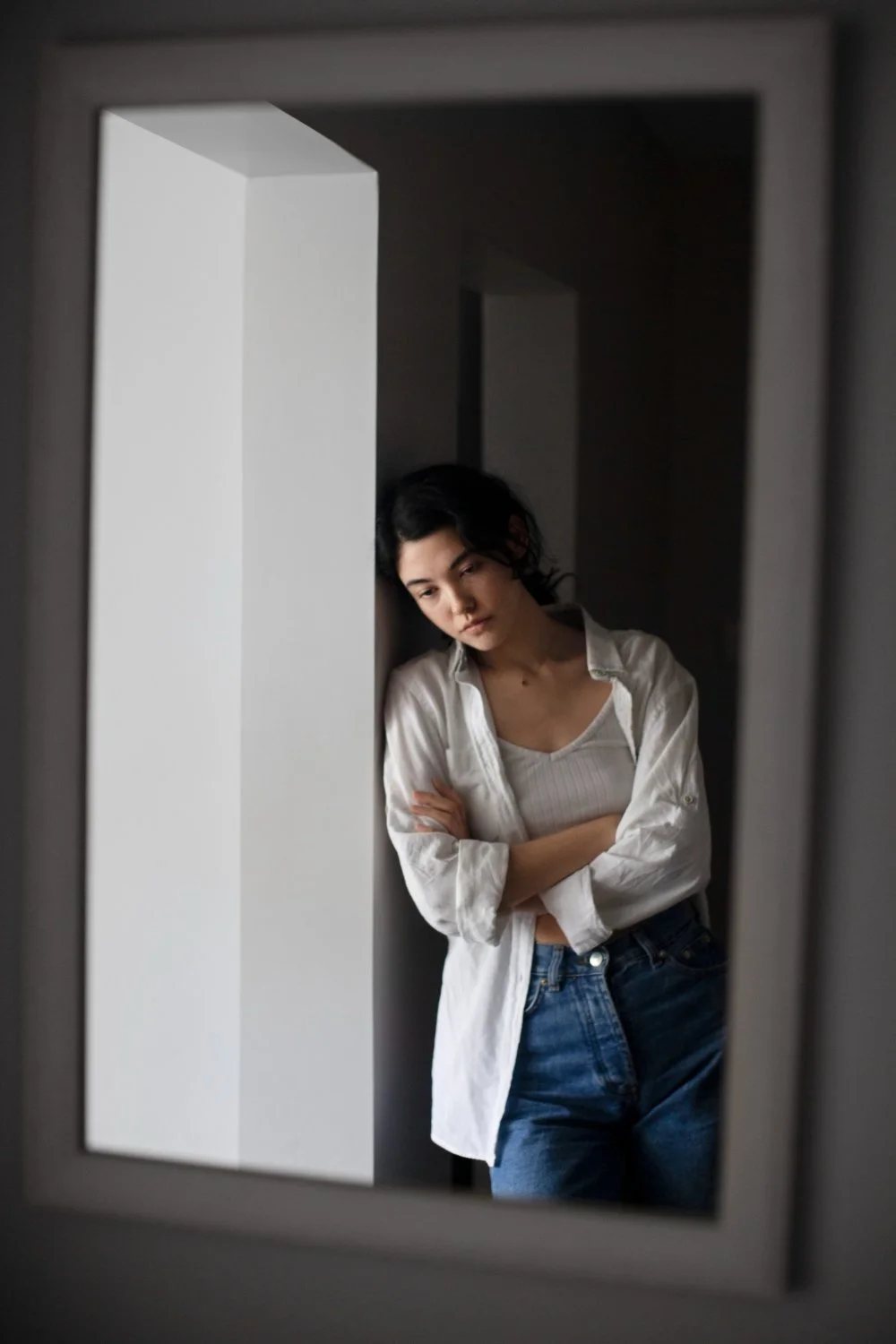 A woman with dark hair, wearing a white shirt over a white tank top and blue jeans, leaning against a wall, looking down thoughtfully.