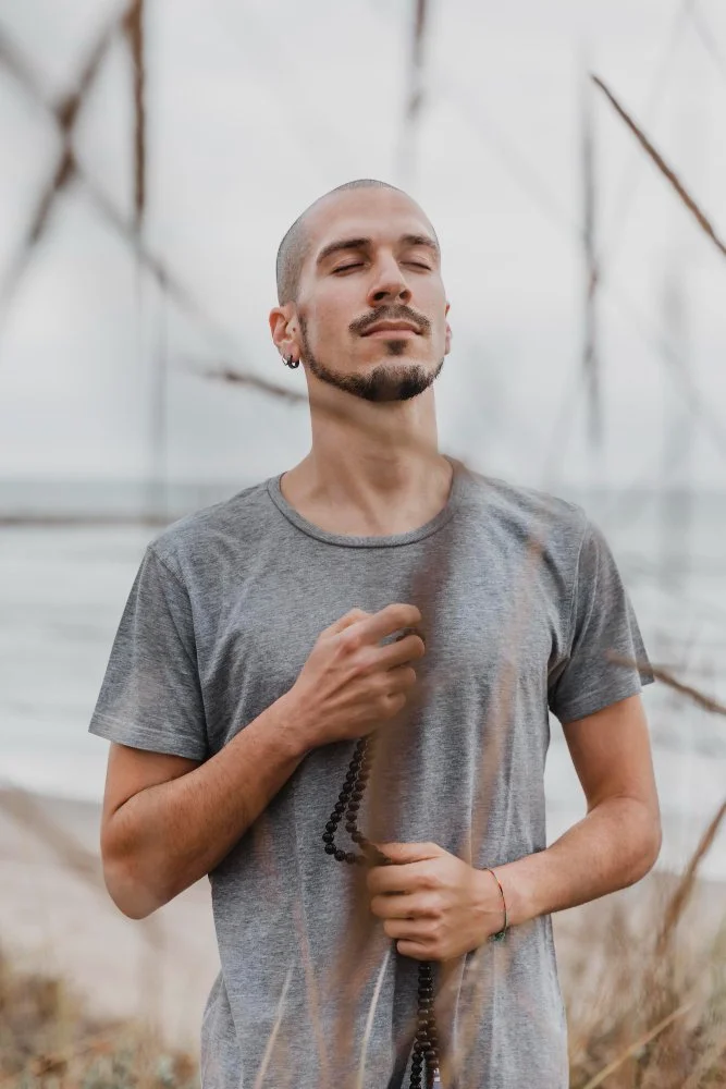 A man with a shaved head, beard, and earrings stands with eyes closed, holding prayer beads, in a natural outdoor setting.