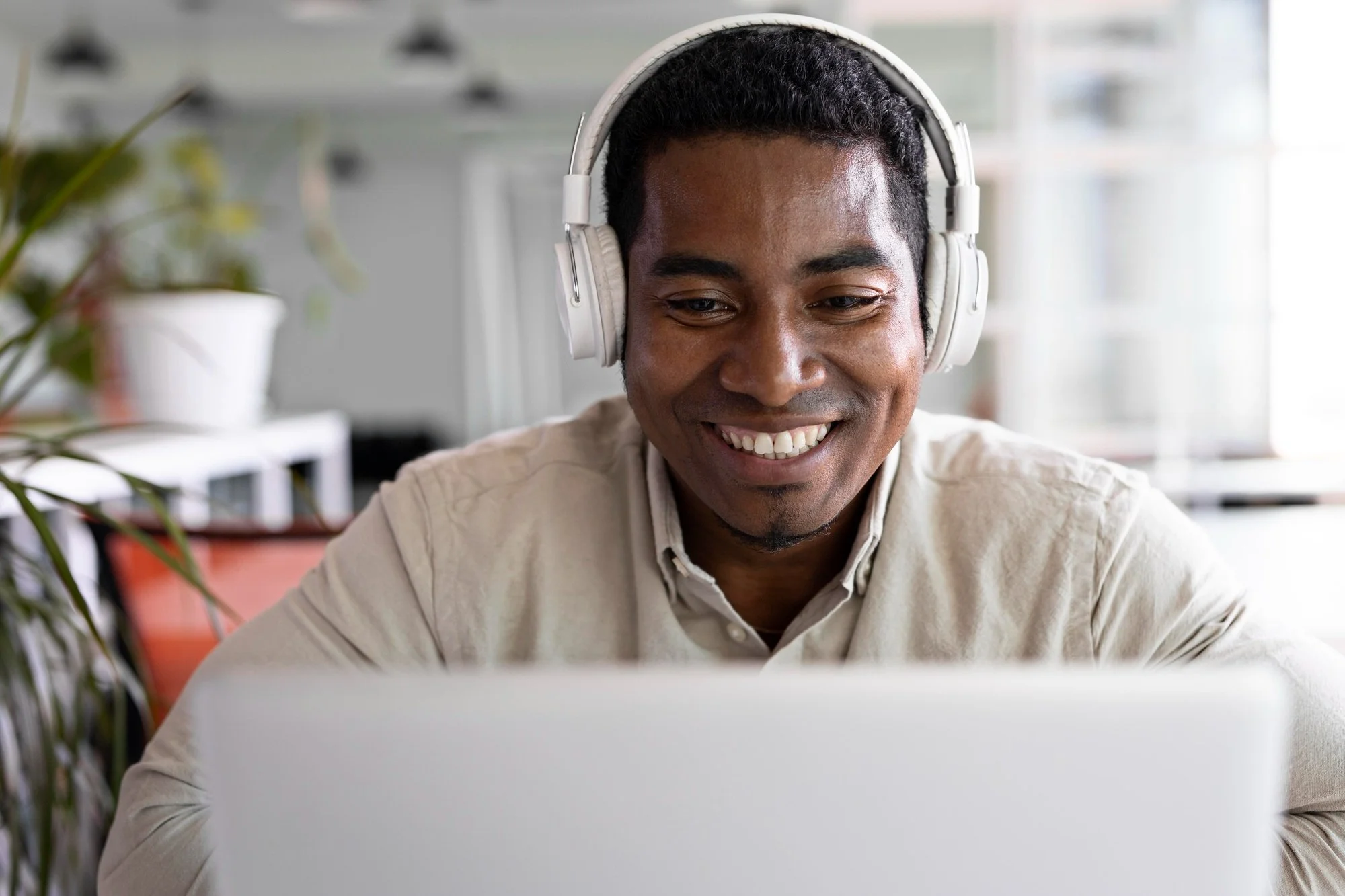 A smiling man with dark curly hair wearing white headphones and a light beige shirt, working on a laptop in a bright, modern office.
