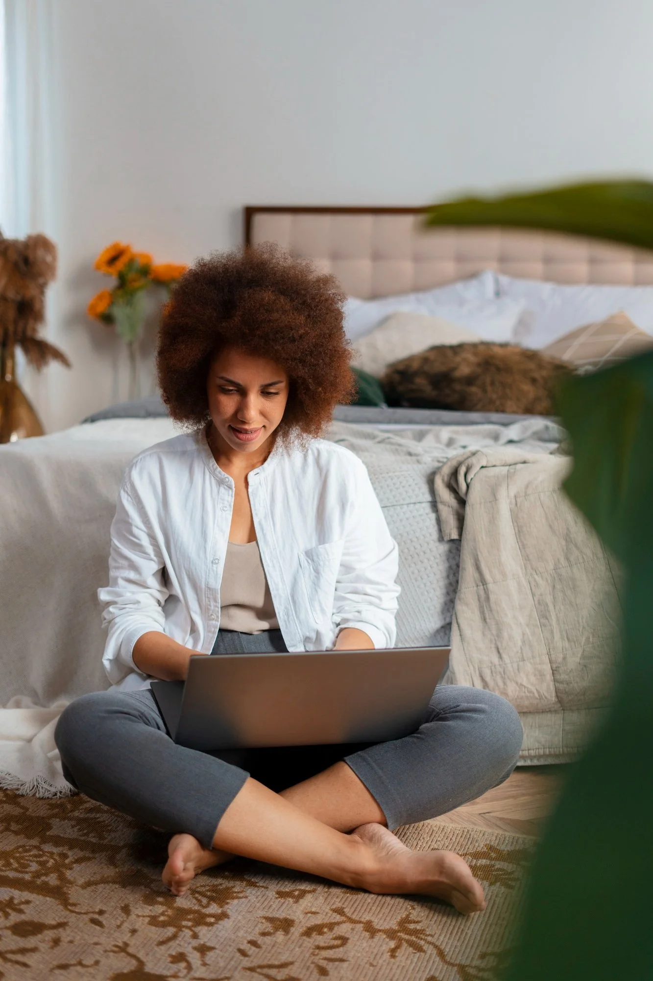 Woman with curly afro hair sitting cross-legged on the floor, using a laptop, indoors with a bed in the background and a blanket draped over a sofa.