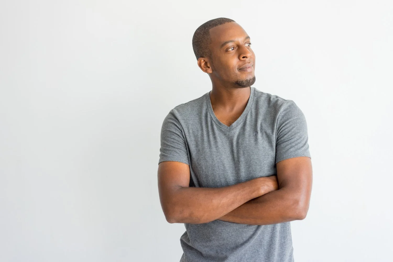 A young man with short hair and a goatee, wearing a gray t-shirt, standing with arms crossed, looking to his right against a plain white background.