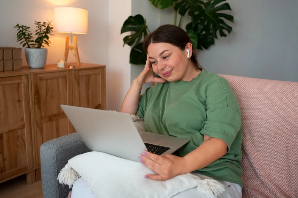 Woman sitting on a pink couch with a laptop, wearing wireless earbuds, in a cozy living room with a wooden cabinet, plants, and a lamp.