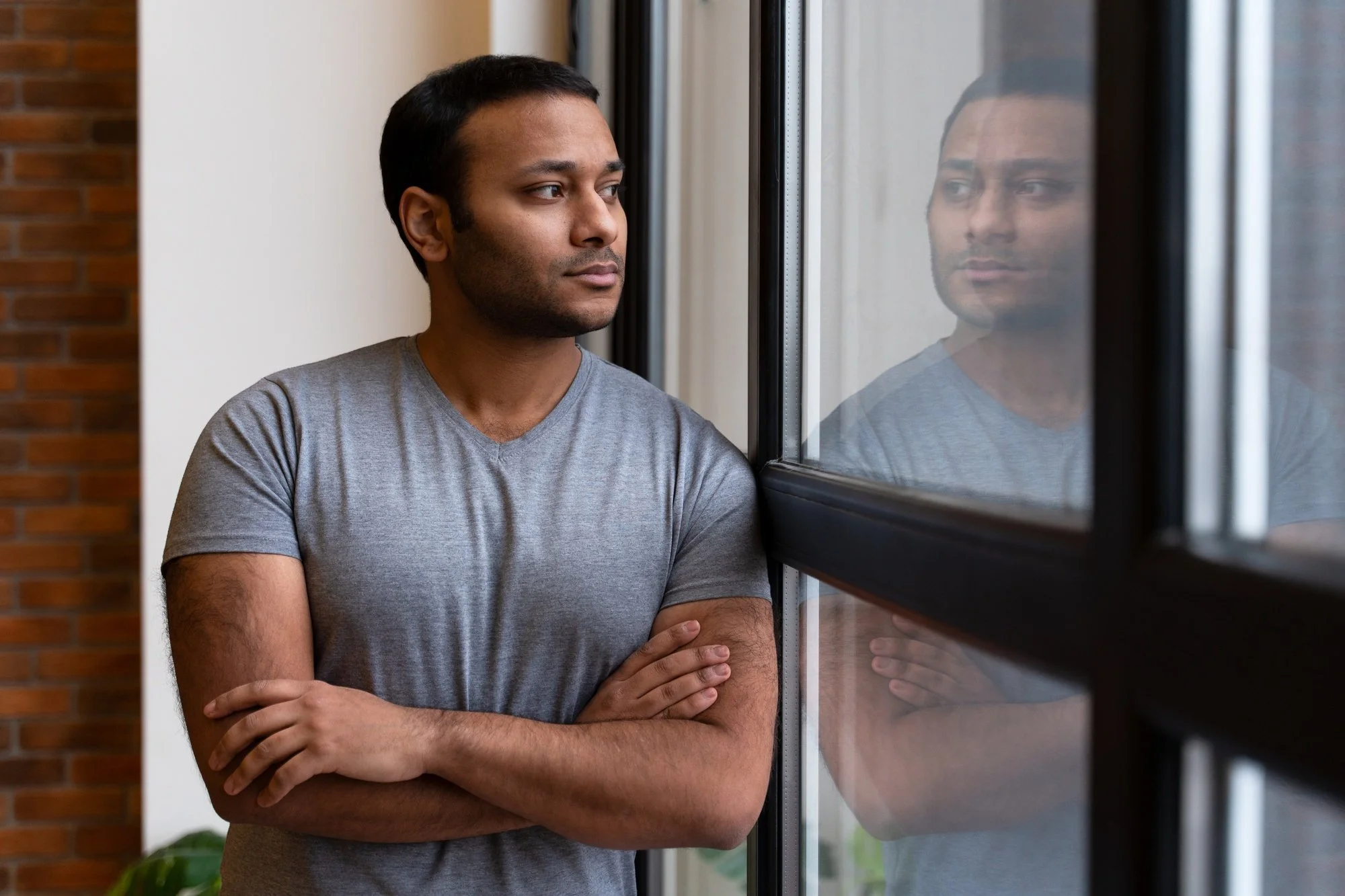 A man with short dark hair and a gray t-shirt standing indoors next to a large window, looking out contemplatively with his arms crossed.