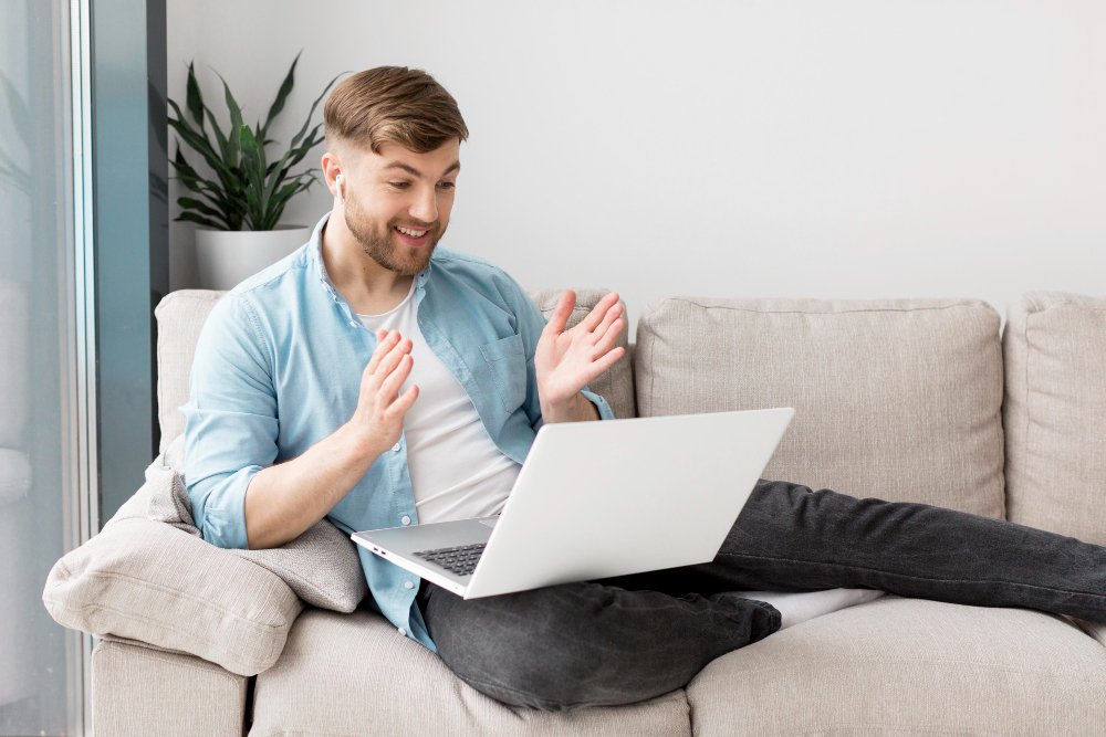 A young man sitting on a beige couch with a laptop on his lap, smiling and gesturing with his hands, in a bright living room with a potted plant nearby.