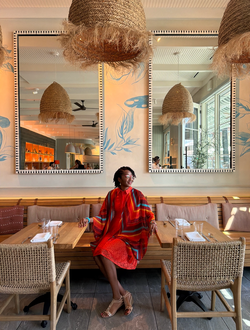 A woman sitting at a restaurant table, smiling, wearing a colorful red and orange dress and beige high heels. The restaurant has wooden tables, woven chairs, decorative hanging light fixtures, mirrors on the wall, and large windows allowing natural light.