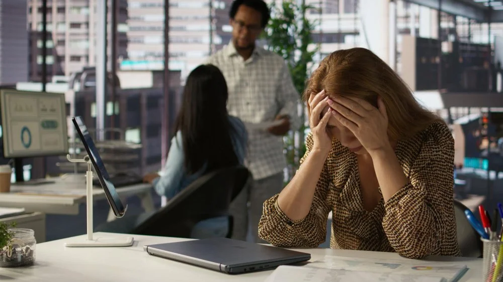 A woman sitting at her desk with her hands on her head, appearing stressed, in a busy office with two colleagues talking in the background.