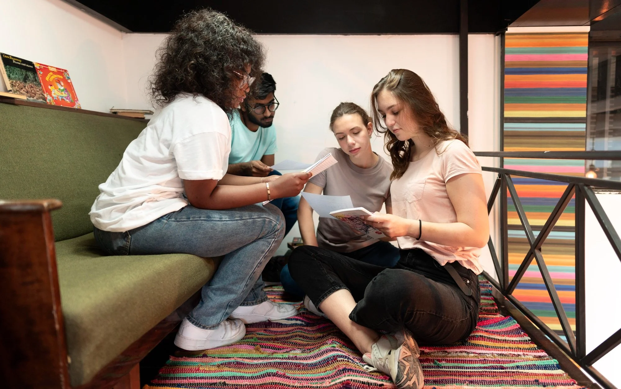 Four young people sitting closely together on the floor, reading printed material, with a bench with books behind them and a colorful striped wall in the background.