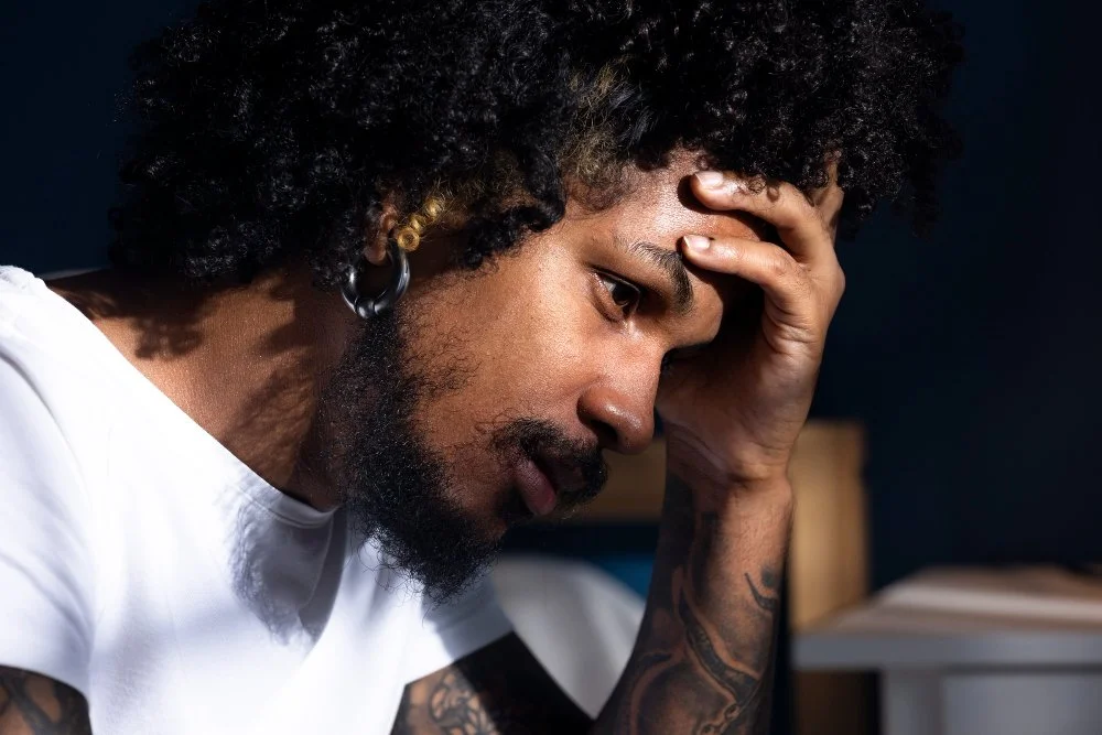 A man with curly hair and a beard, holding his forehead with his hand, appears stressed or worried.