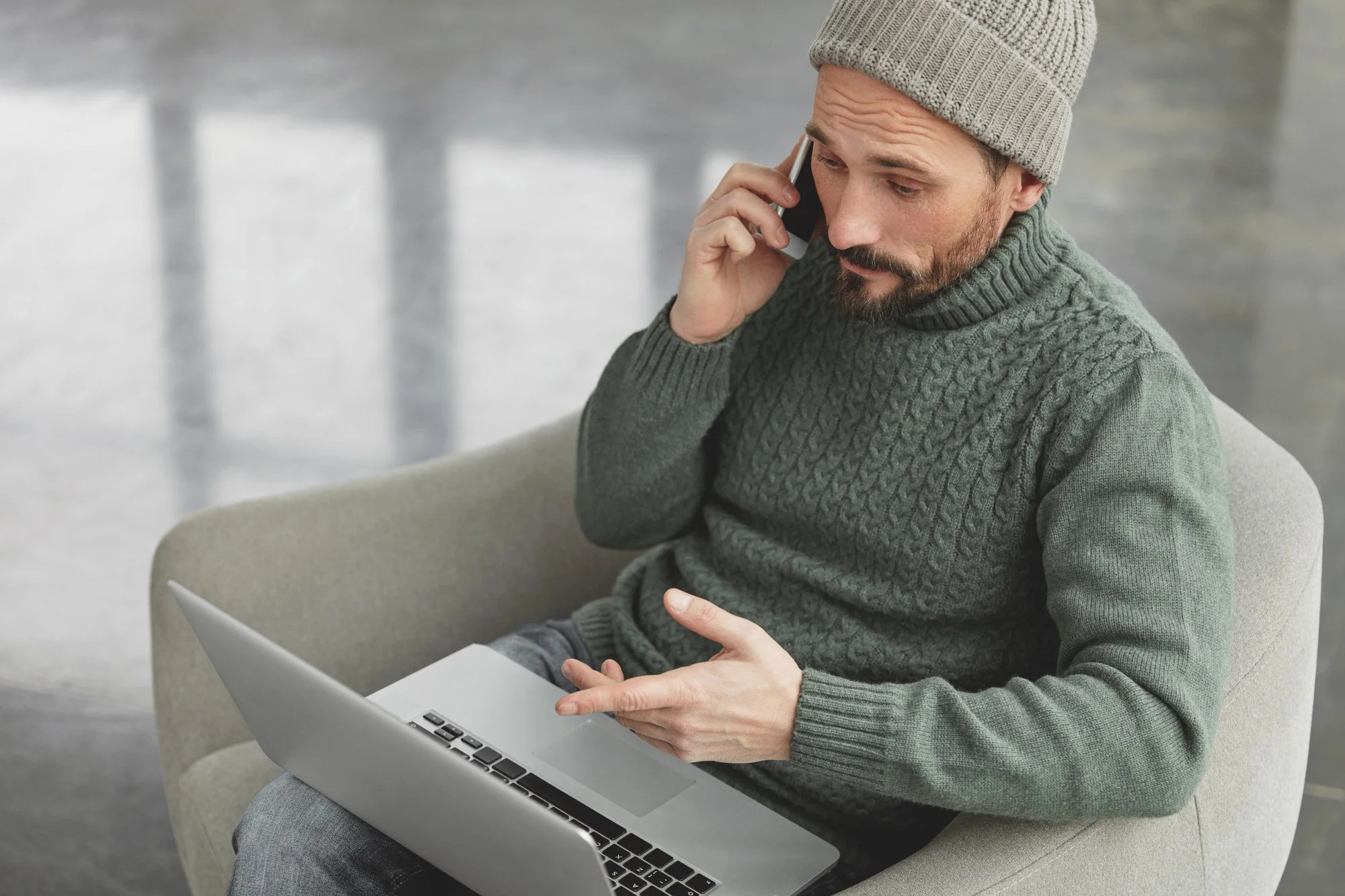 Man in gray sweater and beanie talking on a cell phone while working on a laptop.