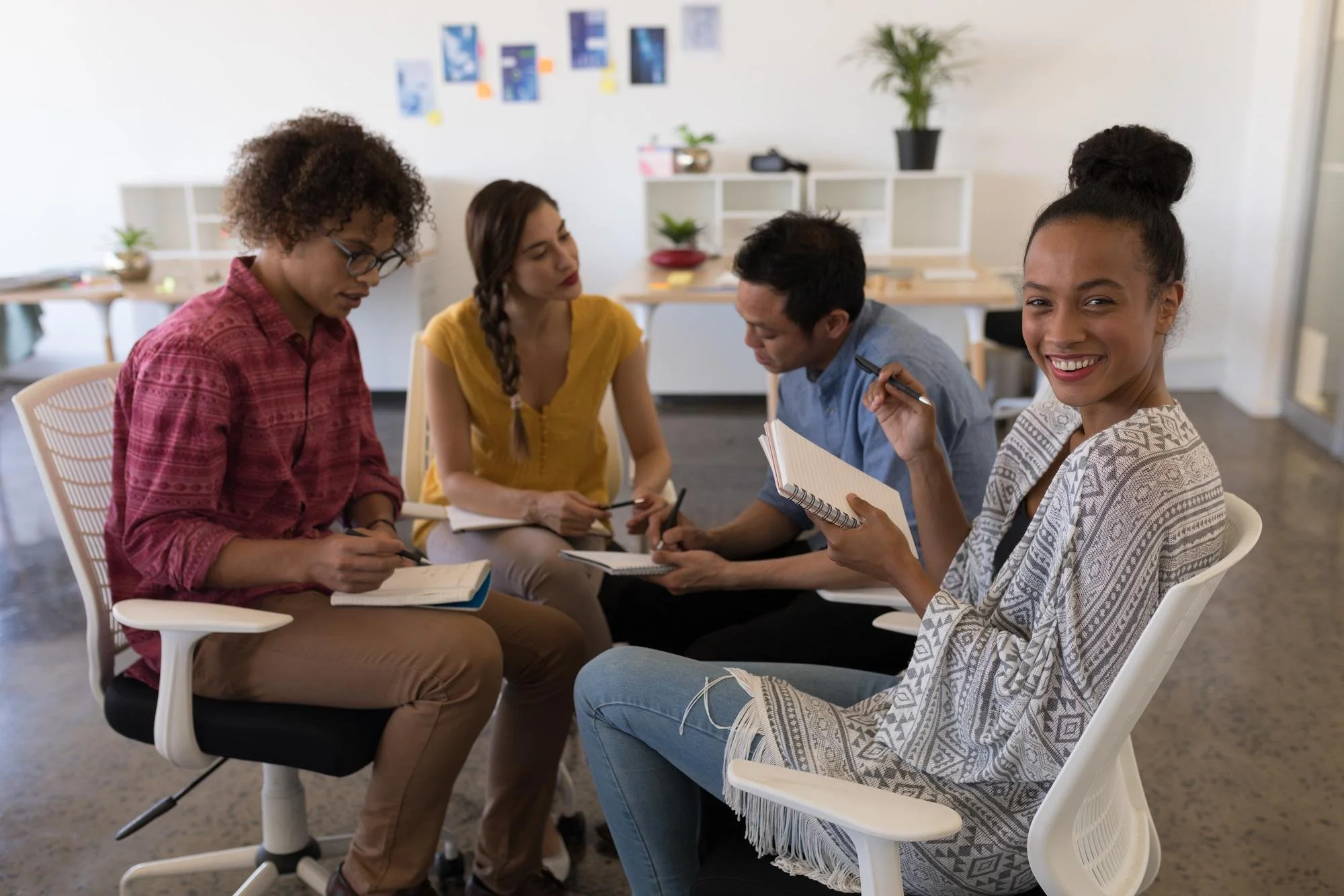 Group of five diverse young adults sitting around a table in a modern office, engaged in a discussion or brainstorming session, with notebooks and pens, smiling and attentive.