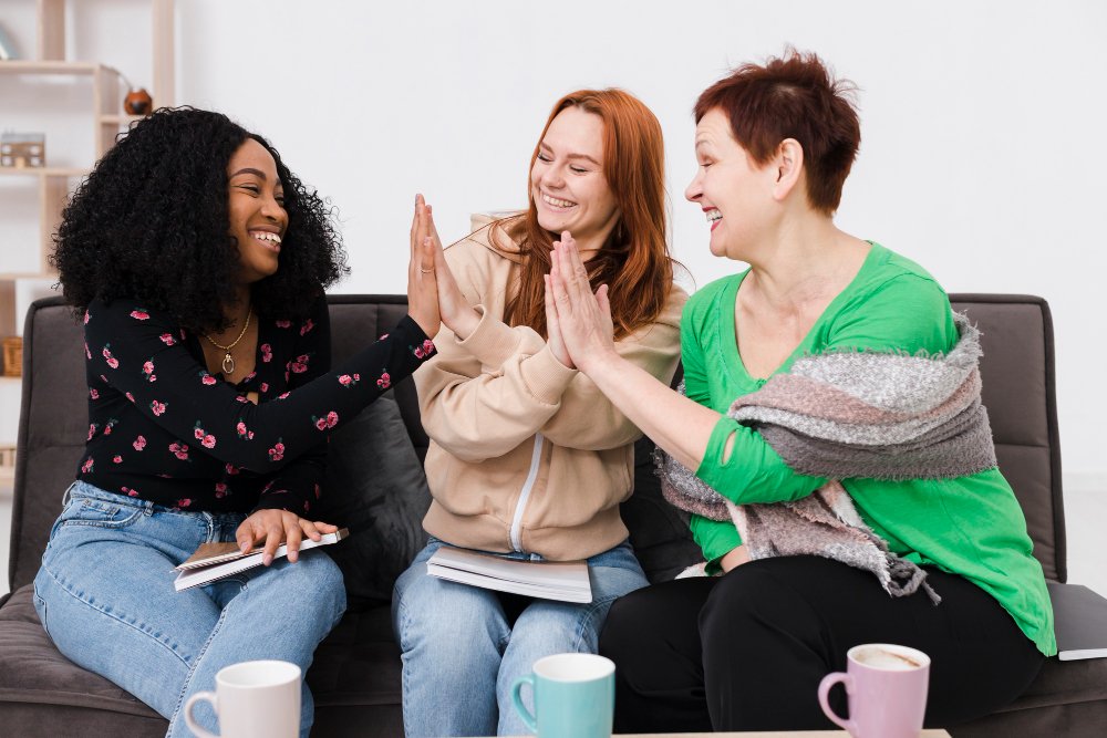 Three women sit on a couch smiling and giving each other high fives in a living room.