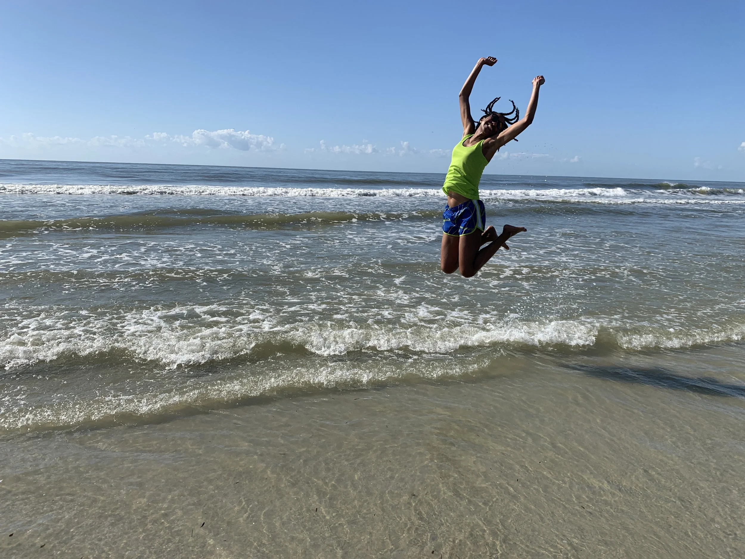 A person jumping in the air at the beach with the ocean and a clear blue sky in the background.