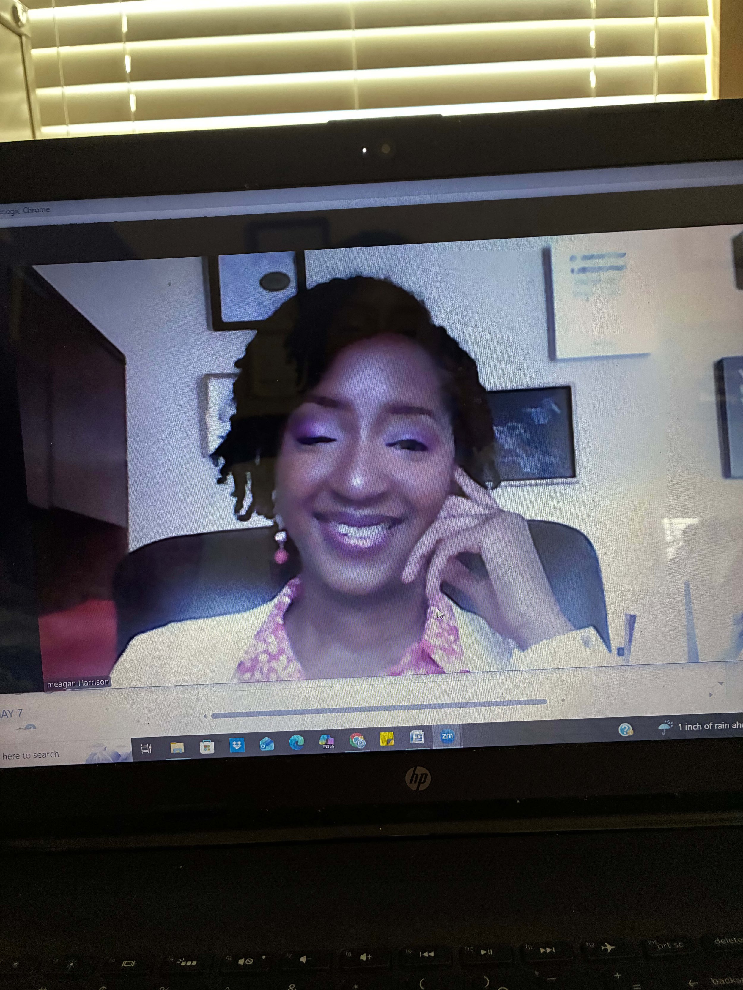 A woman with curly hair and purple makeup smiling and touching her face while on a Zoom video call on a laptop.