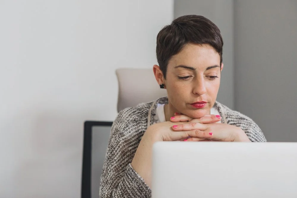 Woman sitting at desk, resting her chin on her folded hands, looking at a laptop.