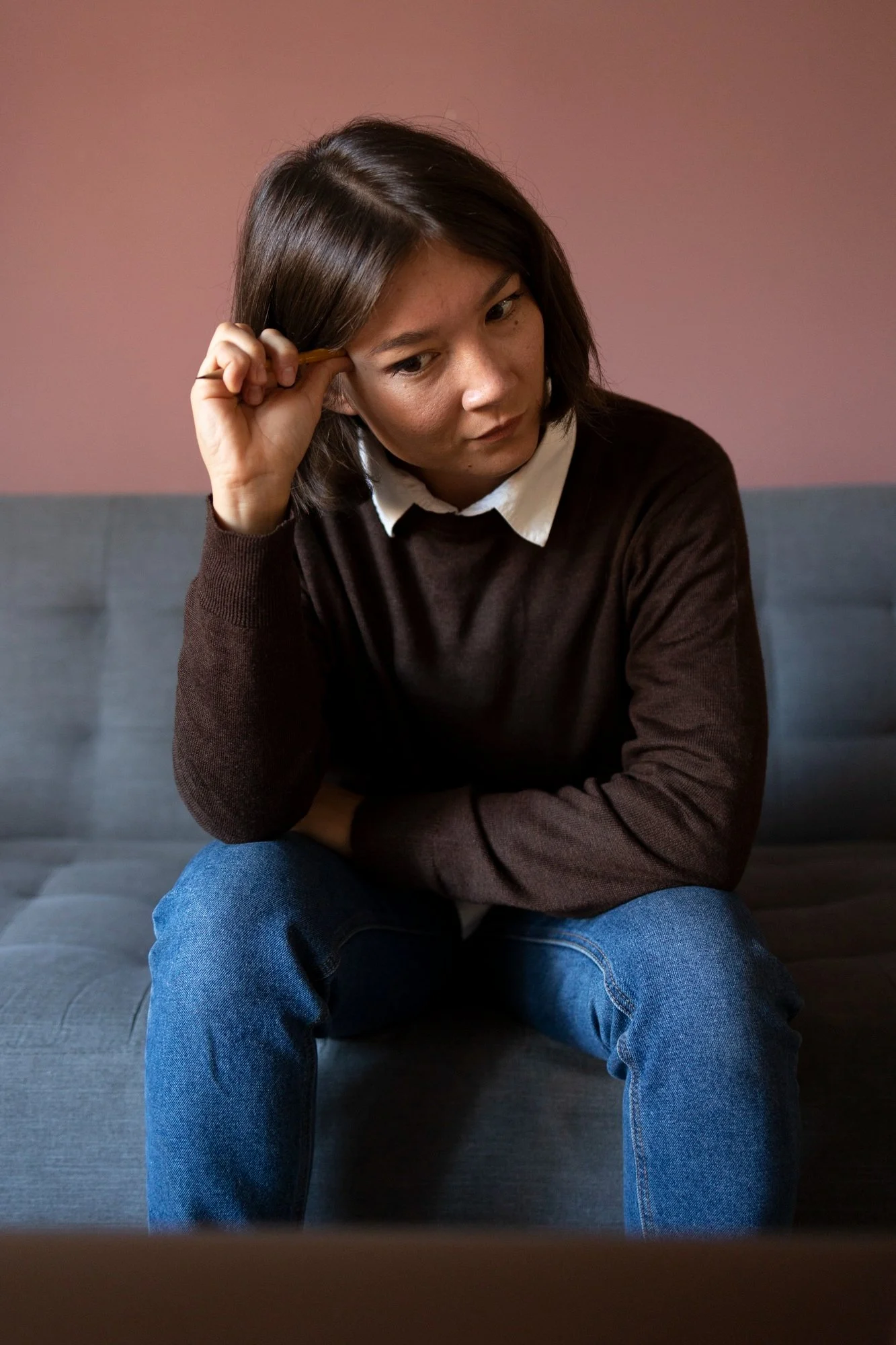 A woman sitting on a gray couch, wearing a white collared shirt and a brown sweater, holding a pen to her temple, looking down thoughtfully with a pink wall in the background.