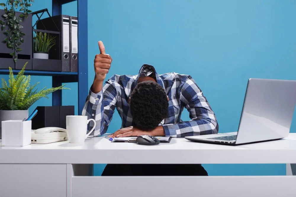 Man with curly hair sitting at a desk with his head down and thumb up, in front of a laptop, with a mug, a pen holder, a desk phone, and a computer mouse on the desk, and books and plants on shelves in the background.