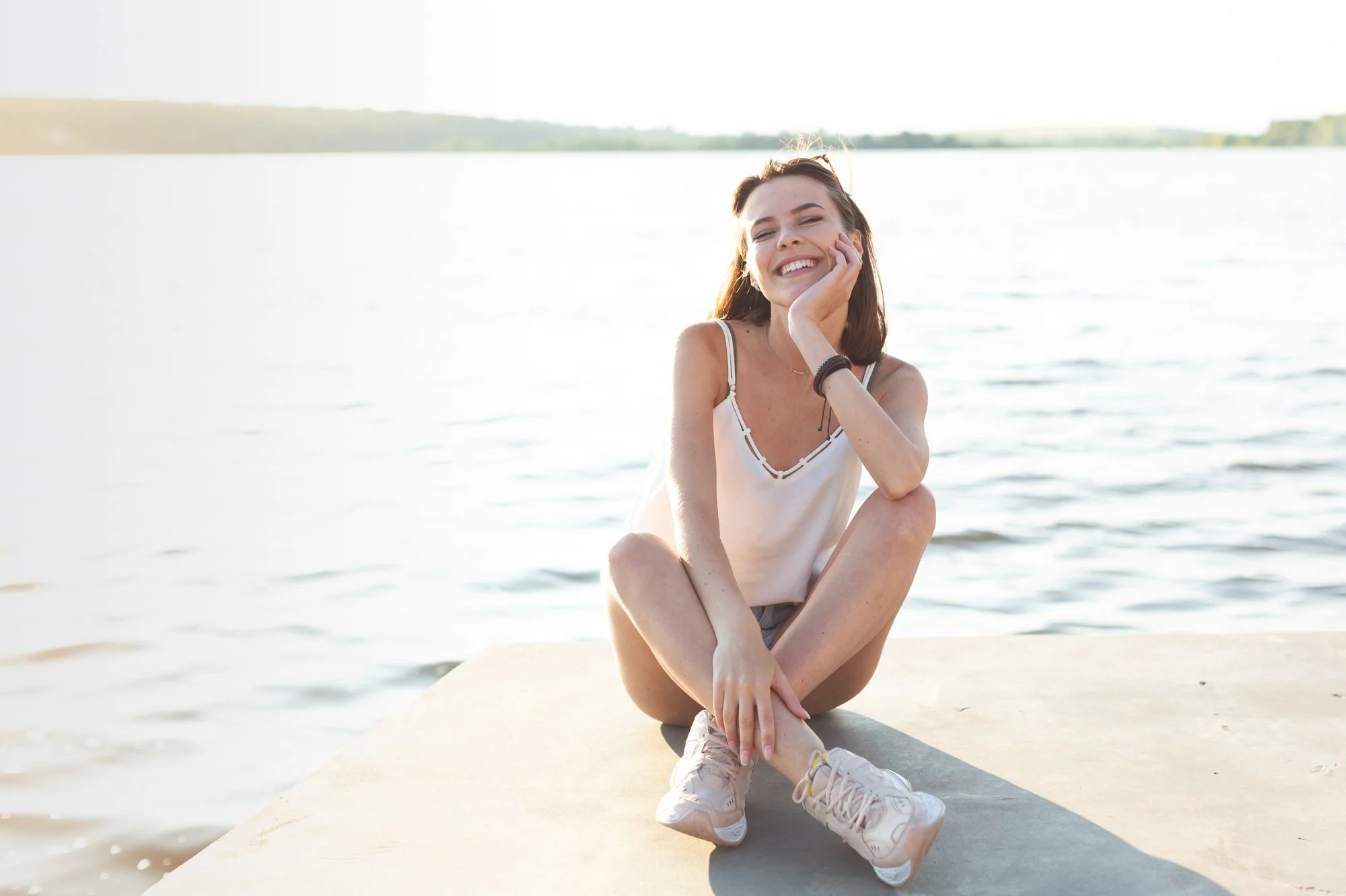 A young woman sitting on a concrete surface by the water, smiling and resting her chin on her hand, with a lake and distant shoreline in the background.